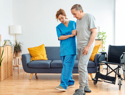 A caregiver assists an elderly man in walking near a wheelchair in a living room with a blue couch and yellow cushions.