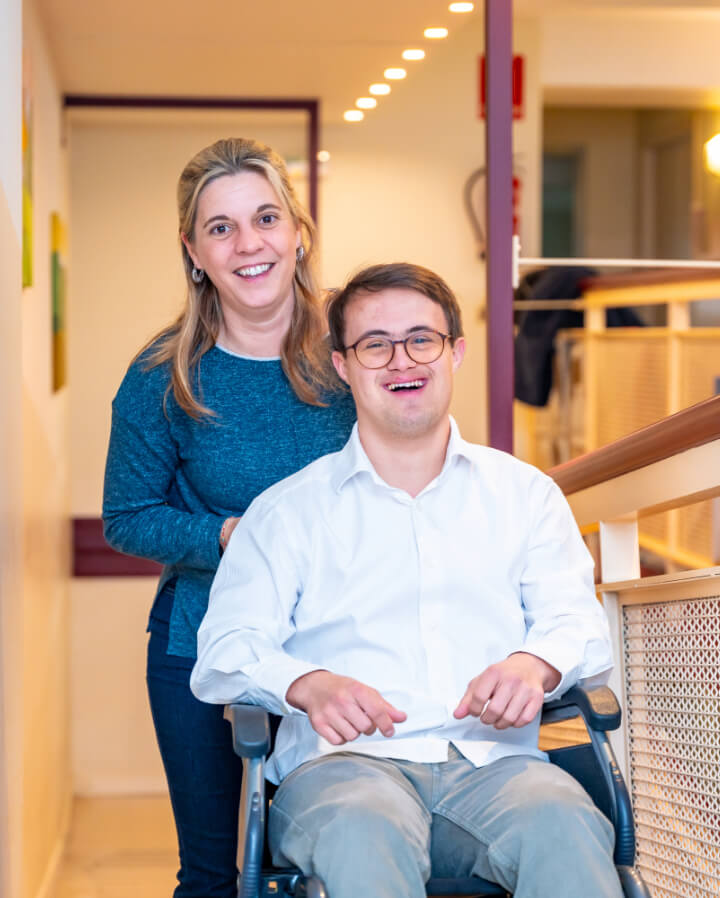 A woman stands behind a smiling man in a wheelchair in a brightly lit indoor setting.